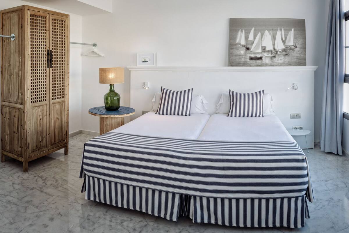 Quarto de hotel com cama grande, guarda-roupa de madeira, mesa lateral redonda, abajur e um quadro na parede.