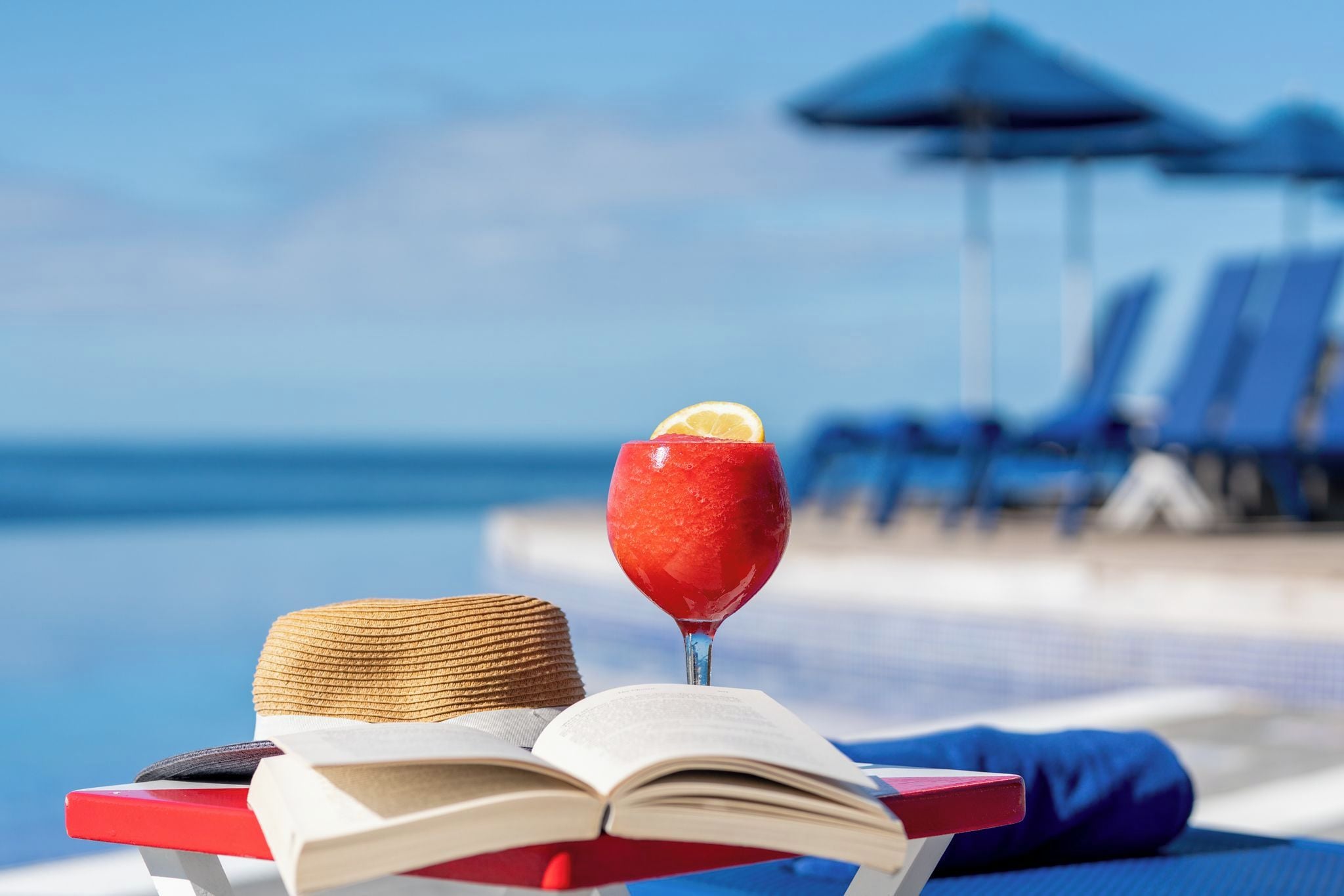 Escena junto a la piscina con un libro, una bebida y un sombrero de paja.