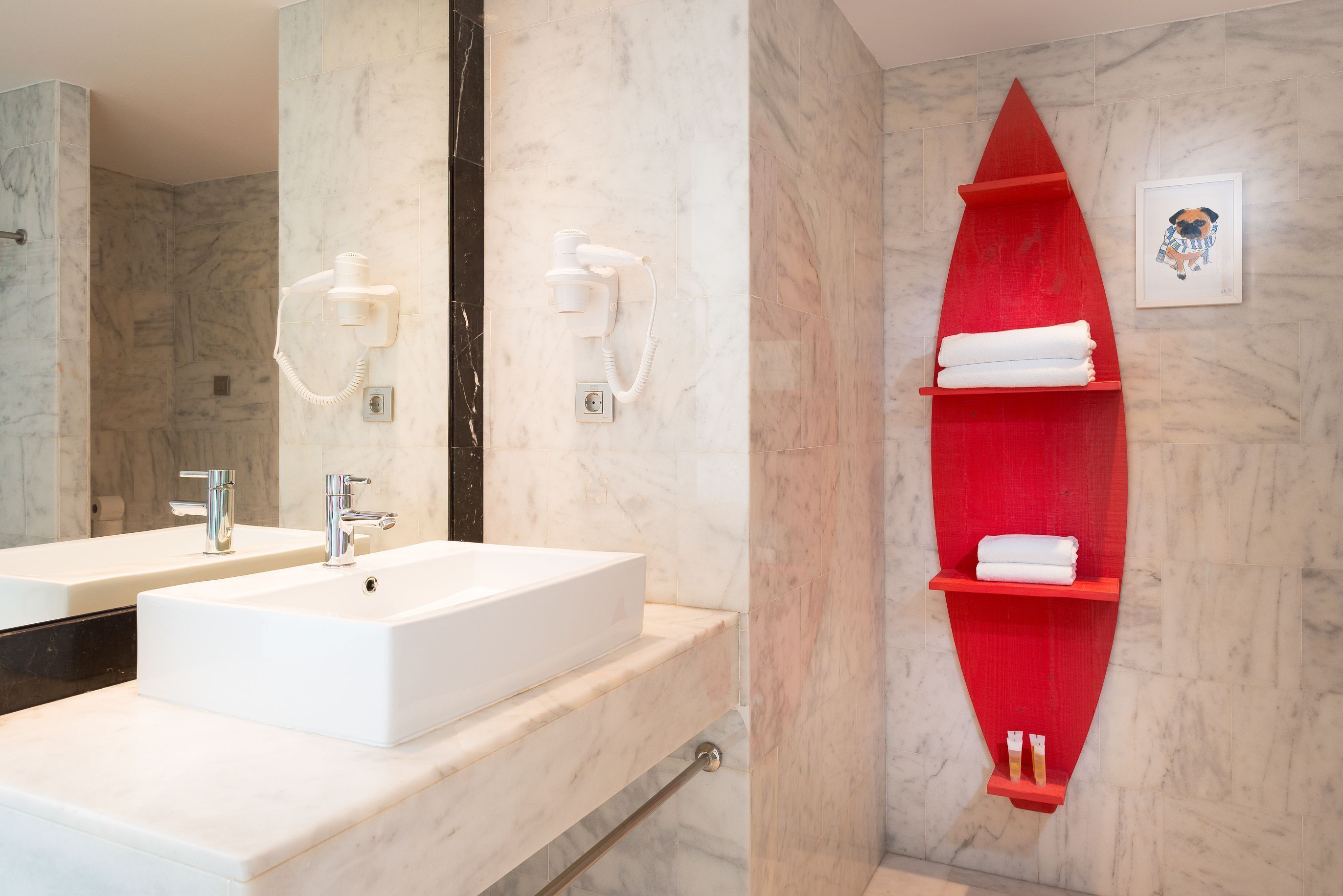 A bathroom with a white sink, two hair dryers mounted on the wall, a red shelf with towels, and a framed picture of a pug.