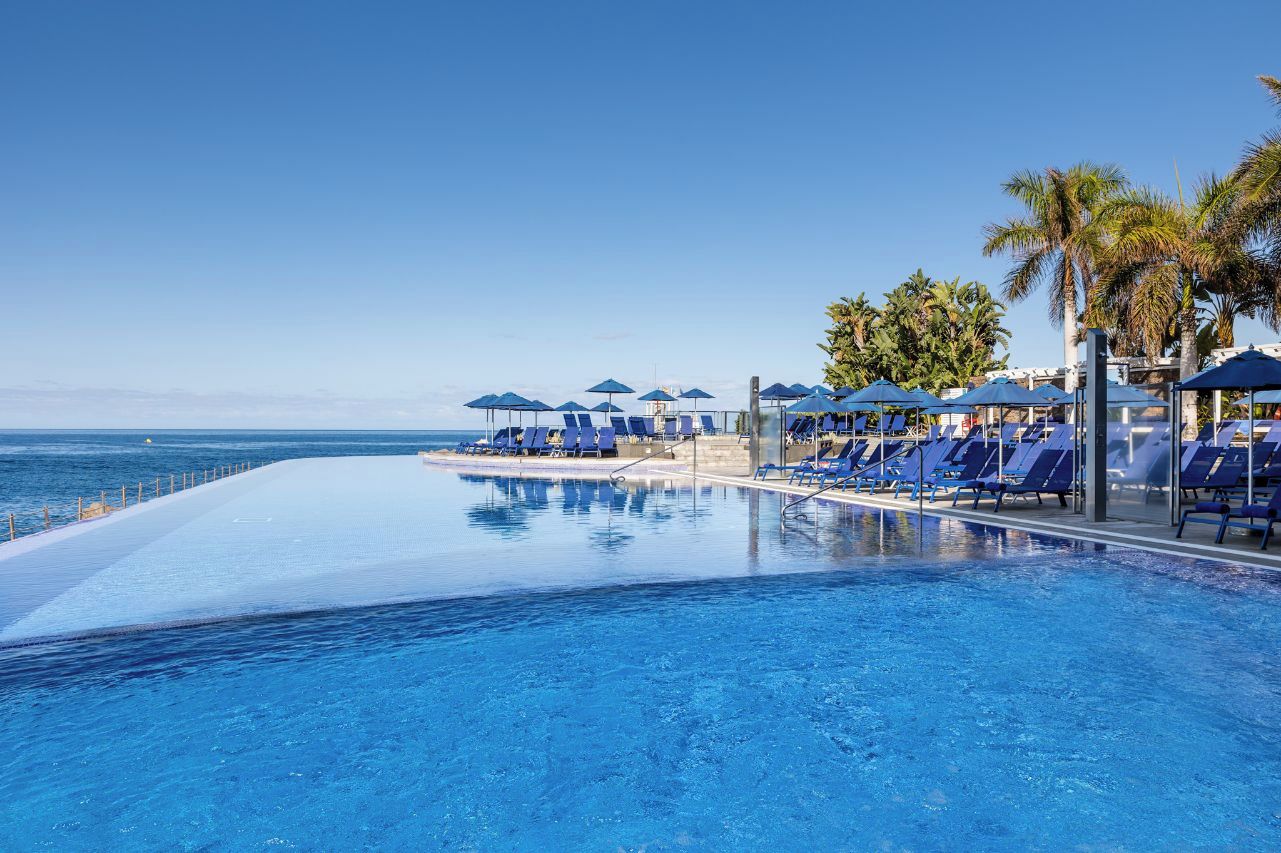 Una piscina con sillones de descanso y sombrillas, cielo despejado y vista al océano.
