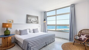 Una habitación de hotel con un ventanal grande, una cama con ropa de cama a rayas, una silla de mimbre y una lámpara sobre una mesa de madera.