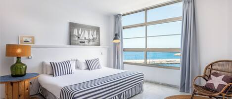 Una habitación de hotel con un ventanal grande, una cama con ropa de cama a rayas, una silla de mimbre y una lámpara sobre una mesa de madera.