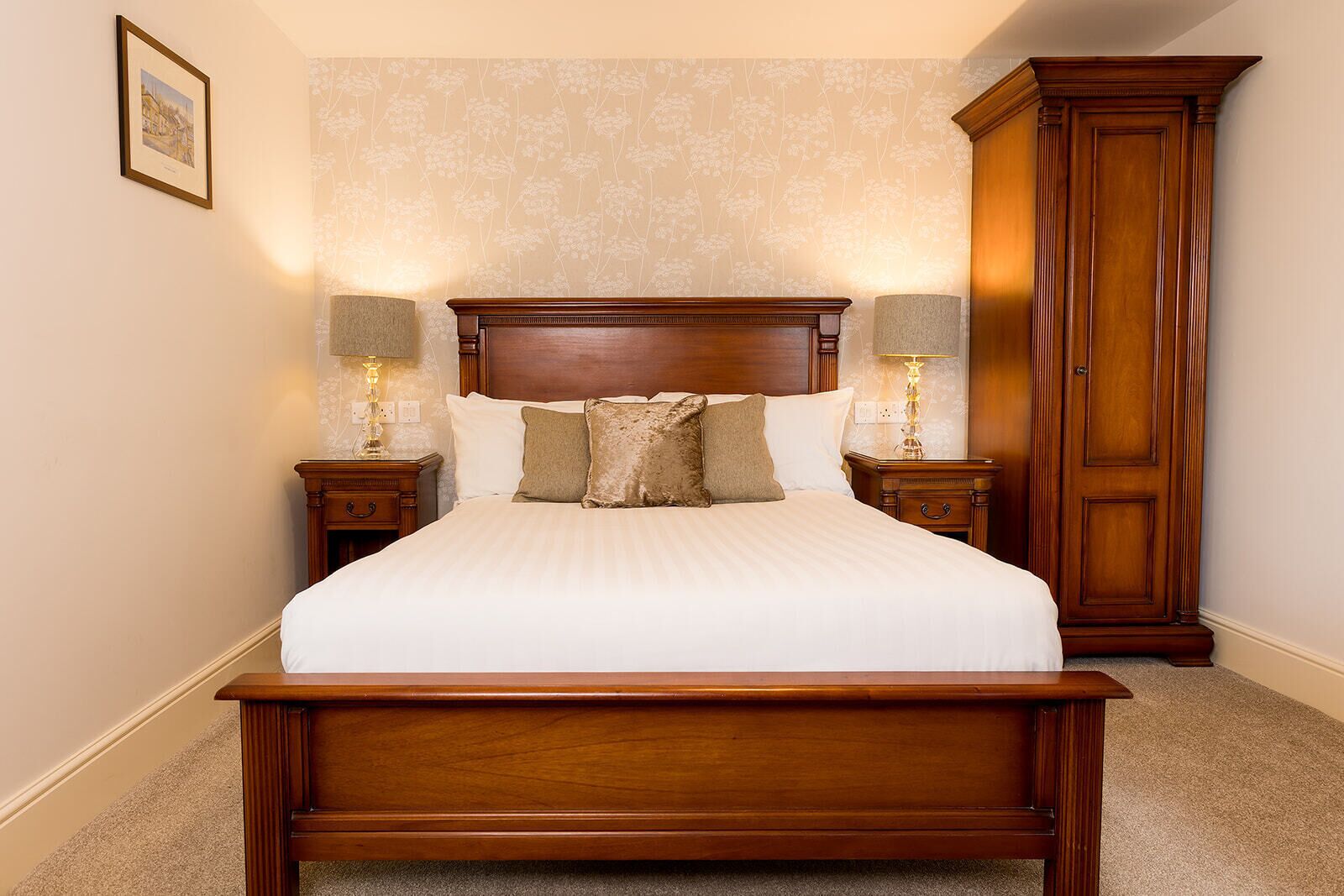 Una cama bien hecha con cabecera de madera, dos mesitas de noche con lámparas y un armario alto de madera.