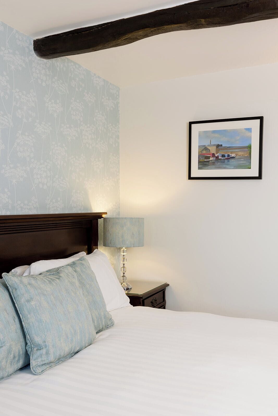 Un dormitorio con cama, almohadas, una mesita de noche con lámpara, un cuadro en la pared y vigas de madera a la vista.