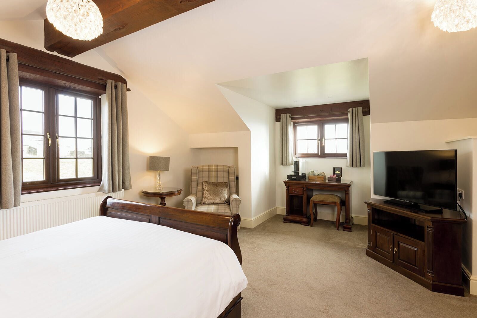 A hotel room with a large bed, a desk with a flat-screen TV, a chair, a lamp, and two windows.
