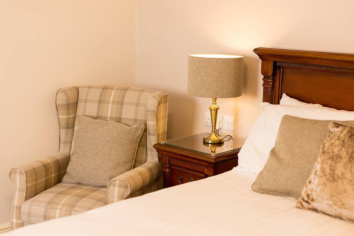 Una camera d'albergo con un letto, una poltrona a quadri, un comodino con una lampada e una testiera in legno.