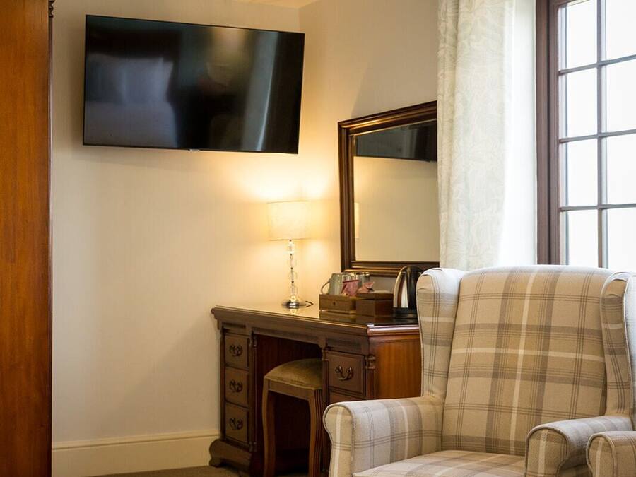 A room with a plaid armchair, a wooden desk with drawers, a flat-screen TV mounted on the wall, and a window with curtains.