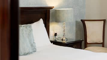 Una cama bien hecha con sábanas blancas, cabecera de madera, una lámpara de noche y una silla con cojín.