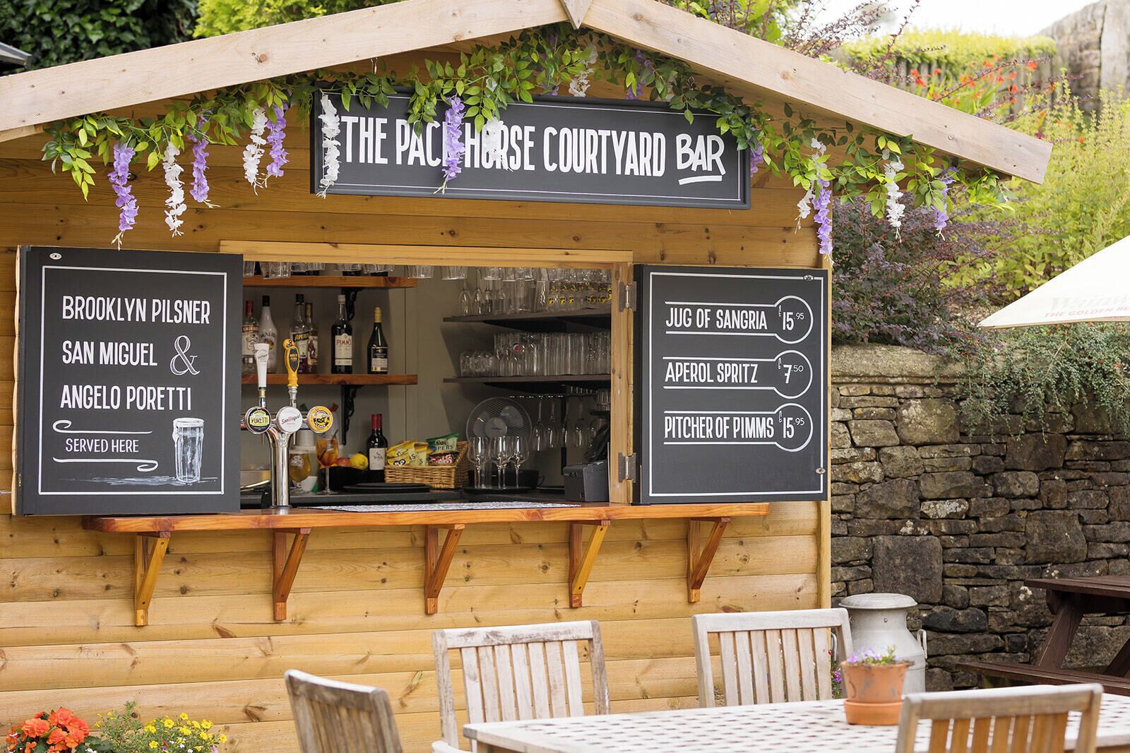 A wooden outdoor bar with a sign reading 'The Pacemaker Courtyard Bar,' a menu board listing drinks, and a view of the surrounding greenery.