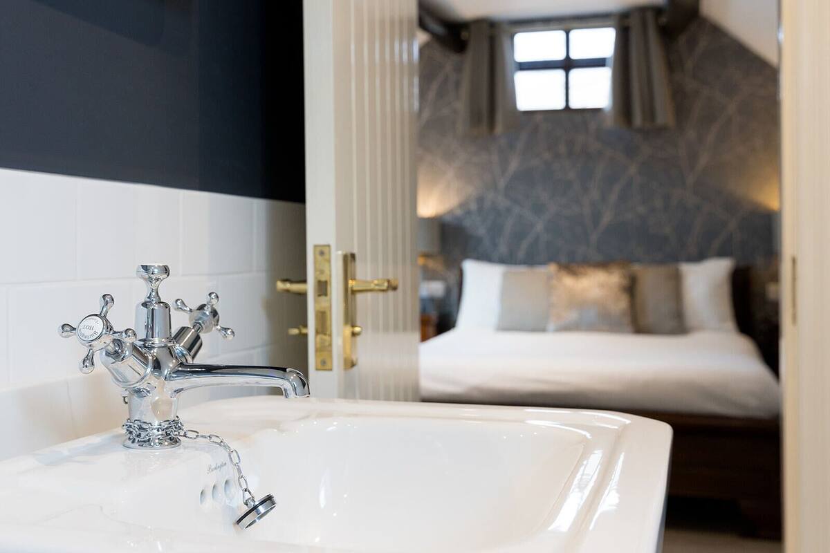 Un bagno con una vasca da bagno ampia e decorata, un lavandino bianco e un ingresso che dà su una camera da letto con un letto e una finestra.
