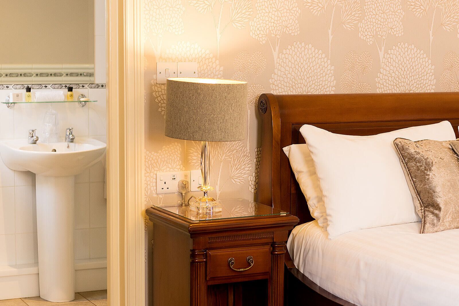 A hotel room with a wooden nightstand, a bedside lamp, a bed with white and brown pillows, and a white sink with a glass shelf above it.