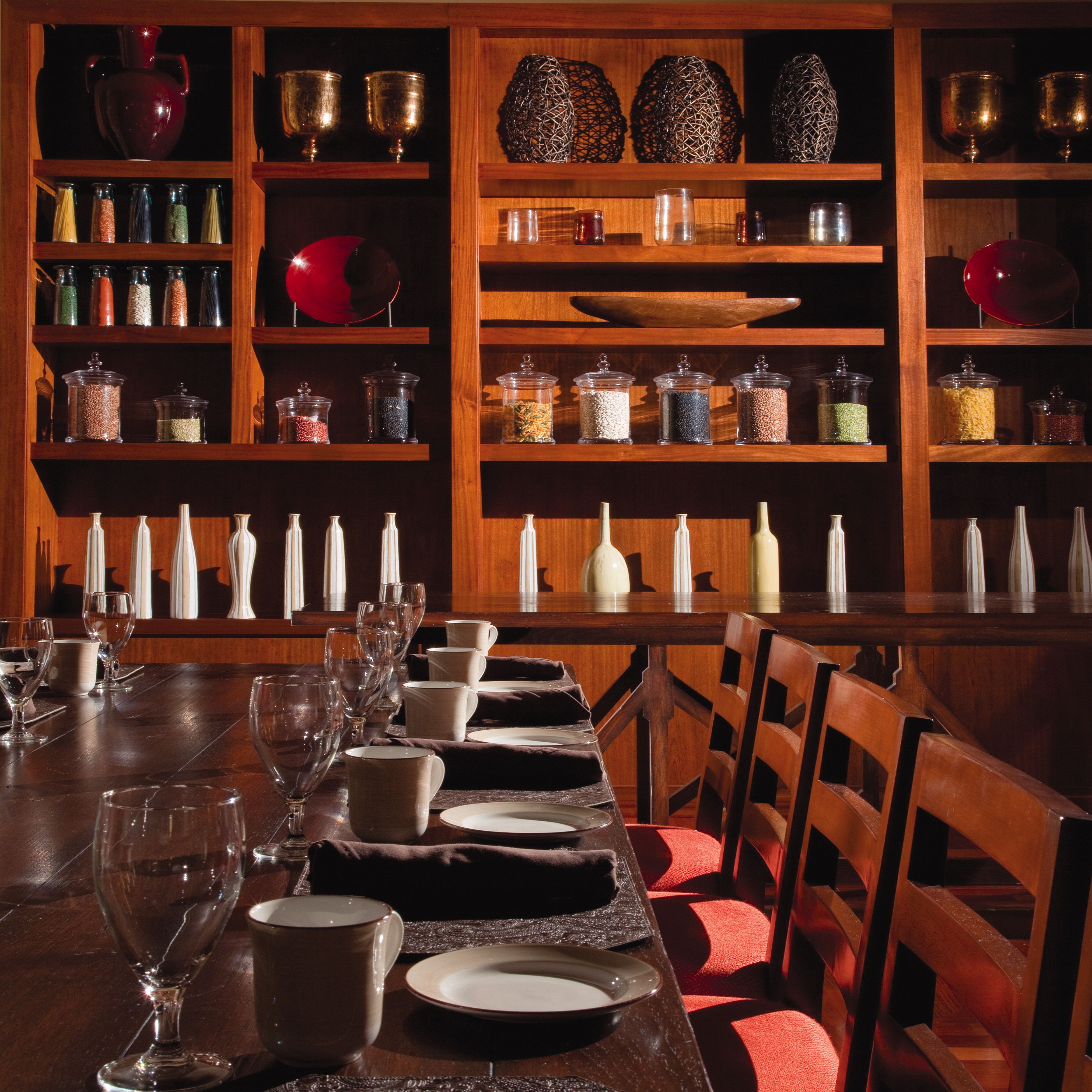 A dining area with a long wooden table set with plates, glasses, and cutlery. Shelves behind the table hold various glass containers and decorative items.