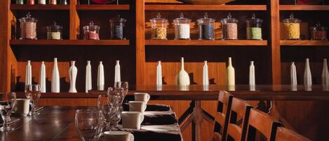 A dining area with a long wooden table set with plates, glasses, and cutlery. Shelves behind the table hold various glass containers and decorative items.