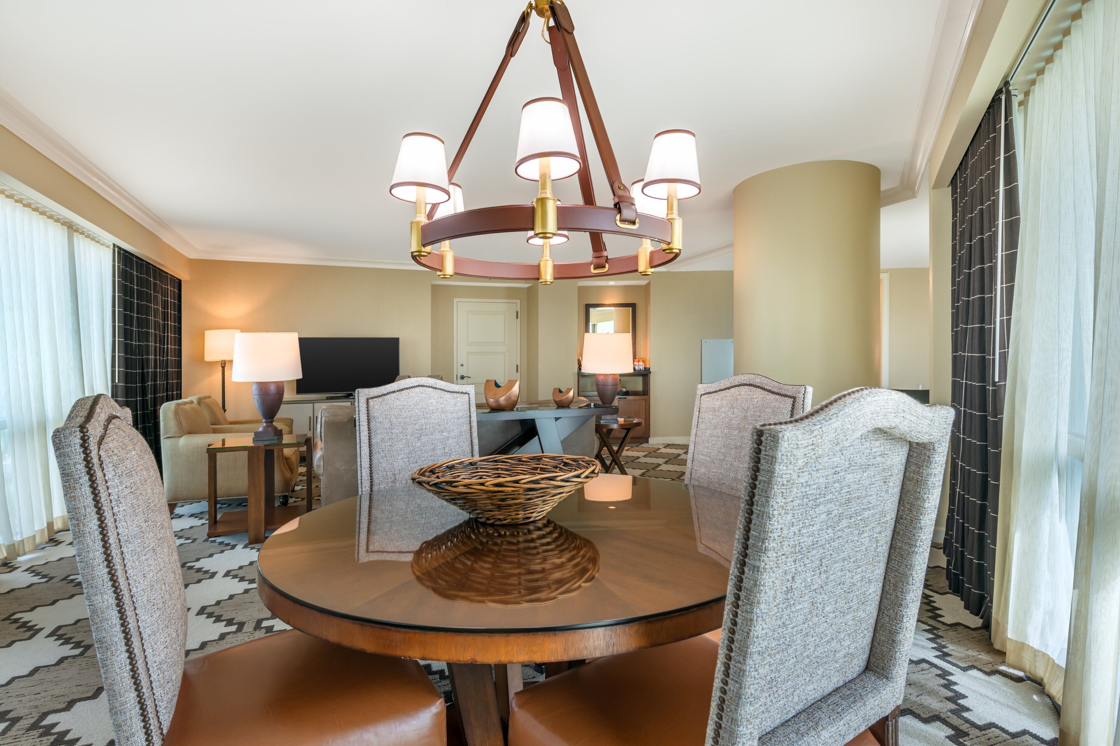 A dining area with a round glass table, leather chairs, a central woven bowl, and a chandelier with multiple lamps.