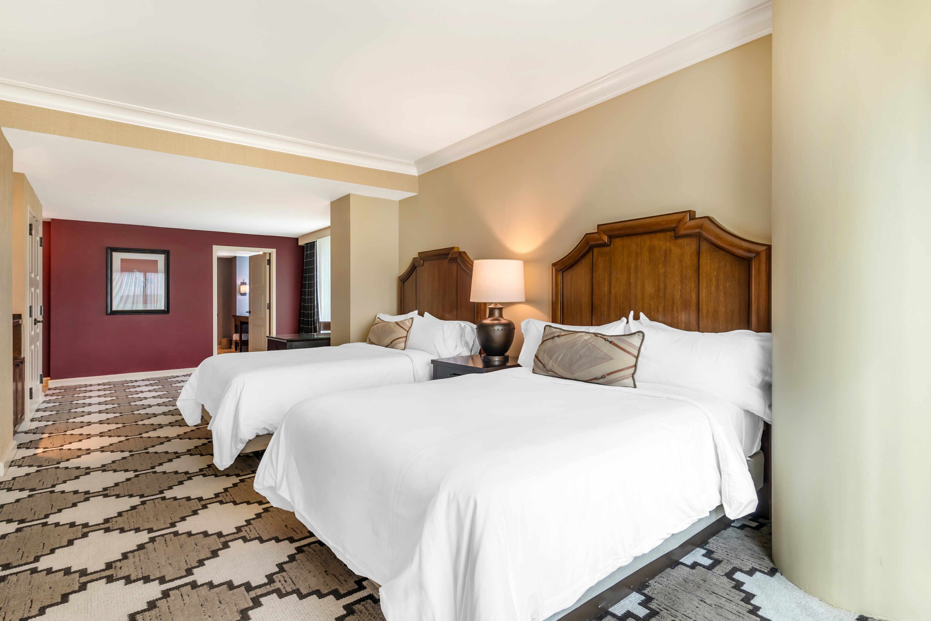 A hotel room with two beds, a patterned carpet, a wooden headboard, and a nightstand with a lamp.