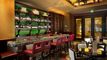 Zona de bar con mostrador de mármol, taburetes con cojines rojos y estanterías llenas de botellas.