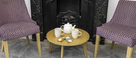 Une pièce avec une cheminée, deux fauteuils à motifs violets, une petite table ronde avec une théière, des tasses et une boîte de thé.