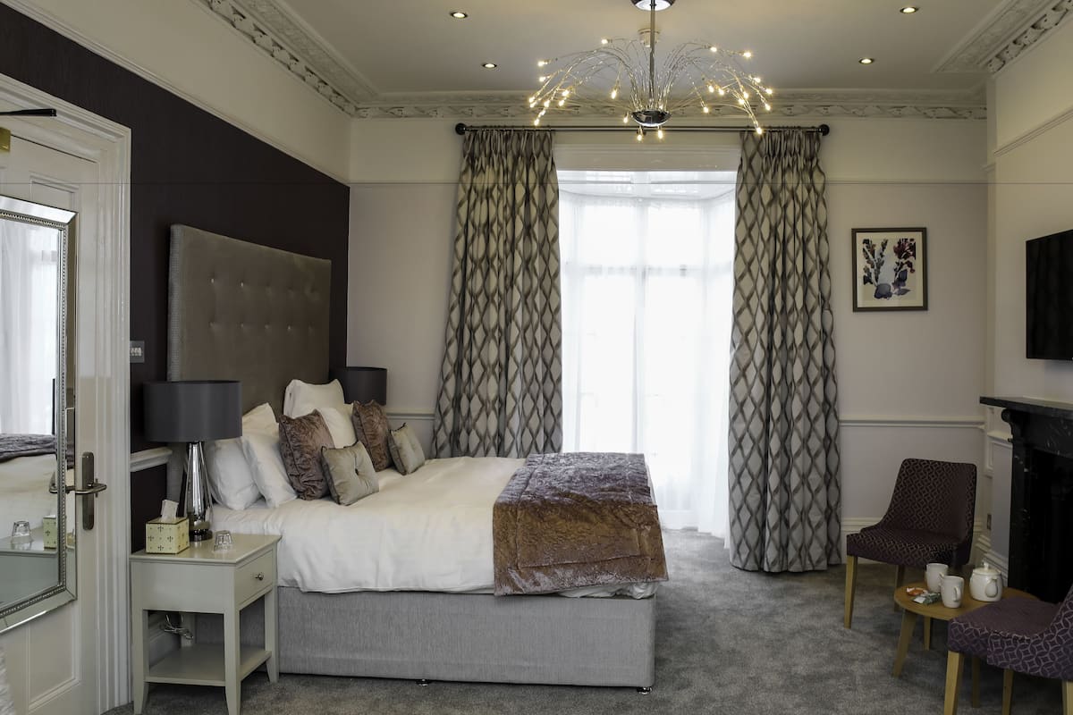A bedroom with a large bed, a bedside table, a chair, a fireplace, and a TV.