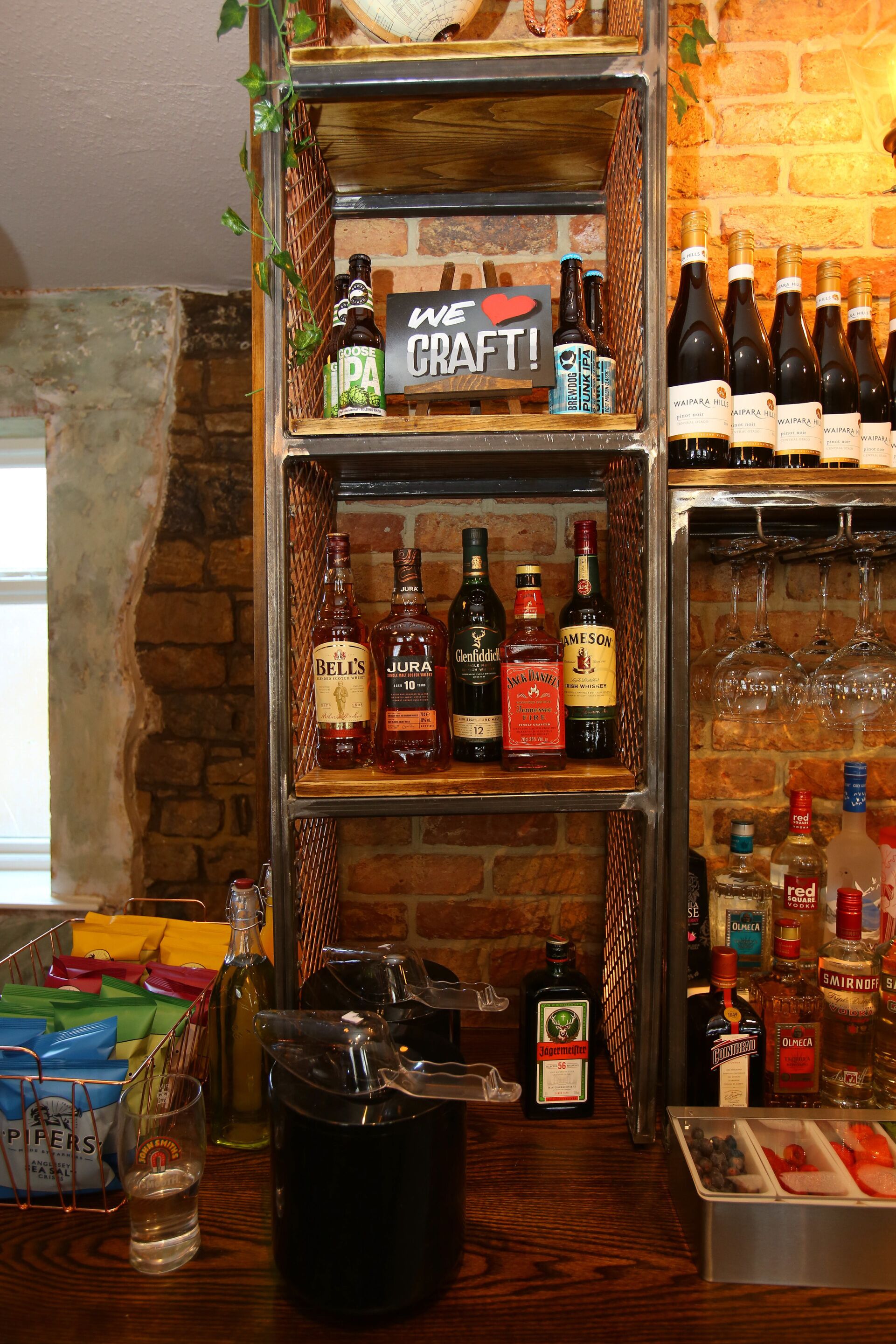 Una zona de bar con estantes llenos de bebidas embotelladas, un cartel que dice 'WE LOVE CRAFT!', y una variedad de vasos y botellas sobre el mostrador.