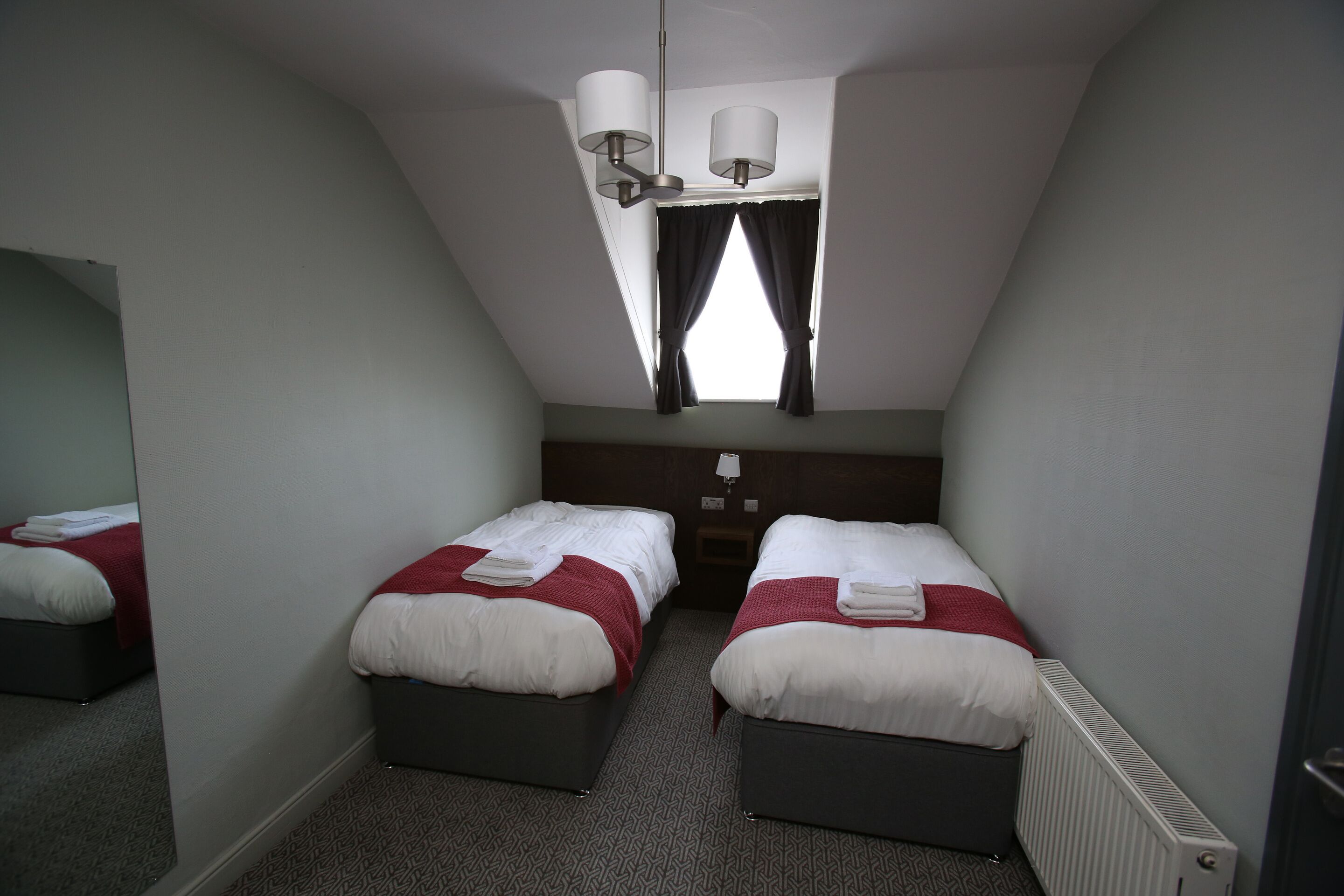 A small, dimly lit room with two beds, a radiator, and a mirror on the left wall.