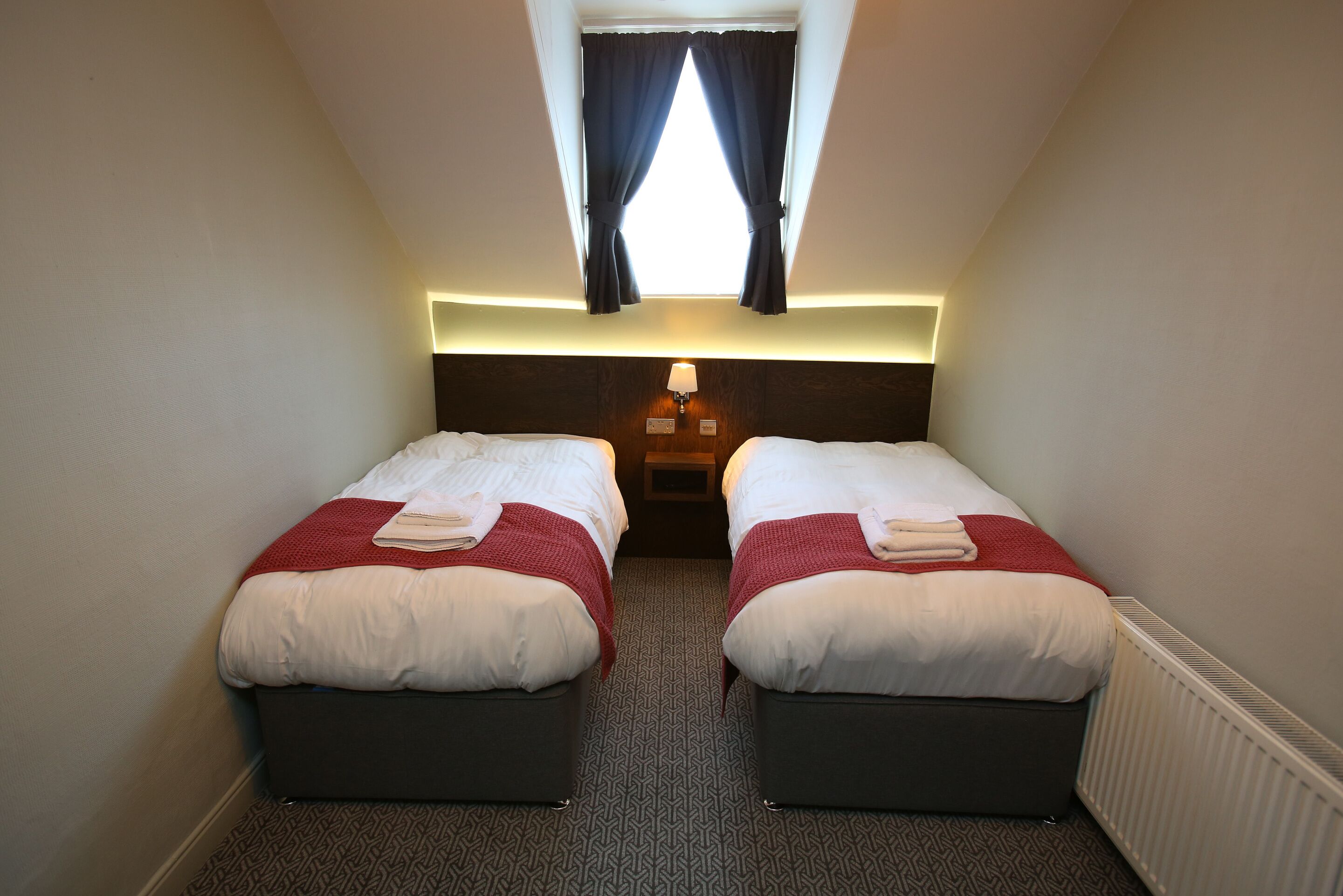 A small, well-lit room with two beds, a nightstand with a lamp, and a window with curtains.