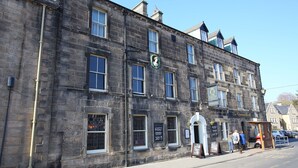 A historic stone building with multiple windows, a sign for 'The Queens Head' pub, and a sidewalk with a dog and people.