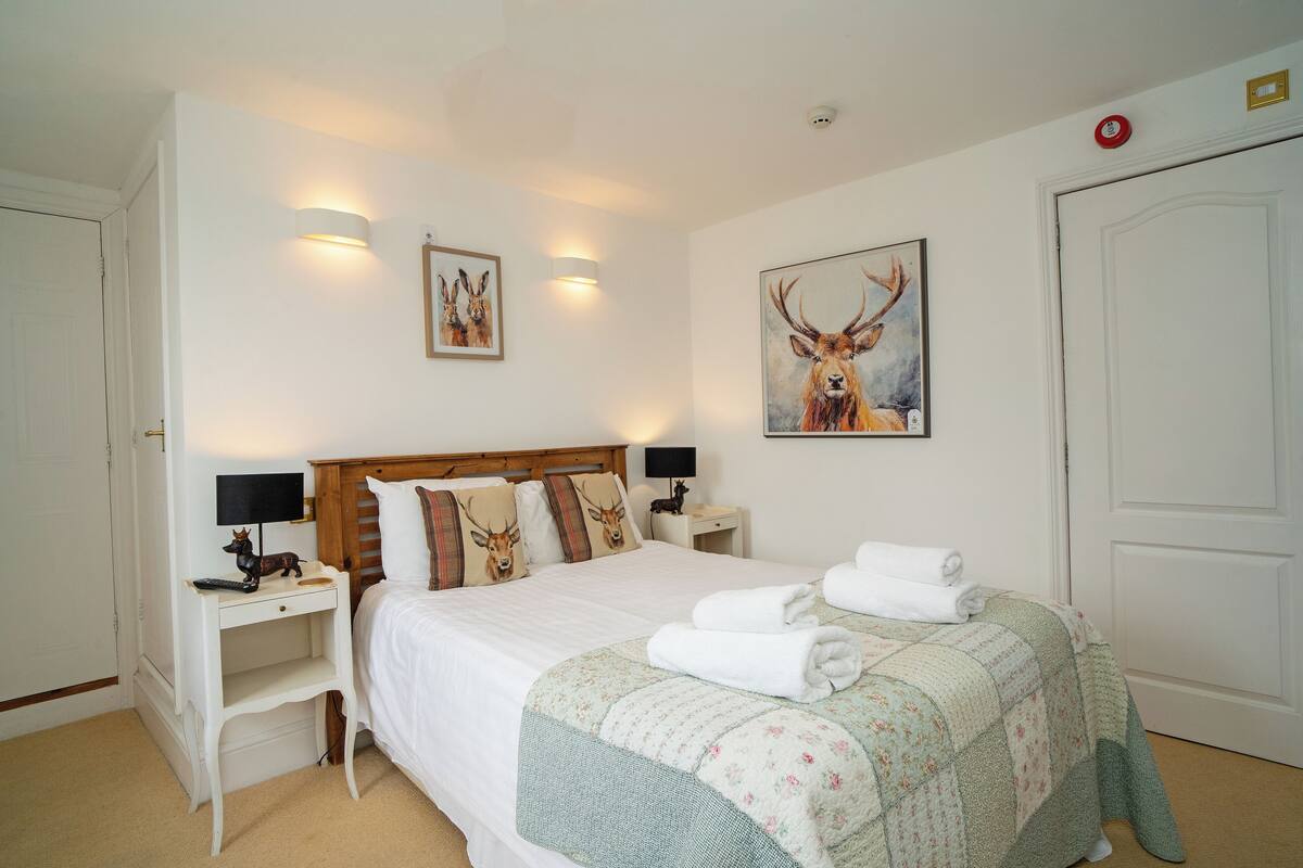 A neatly arranged bedroom with a large bed, two bedside tables with lamps, a wall-mounted artwork of a deer, and a door with a red emergency light.