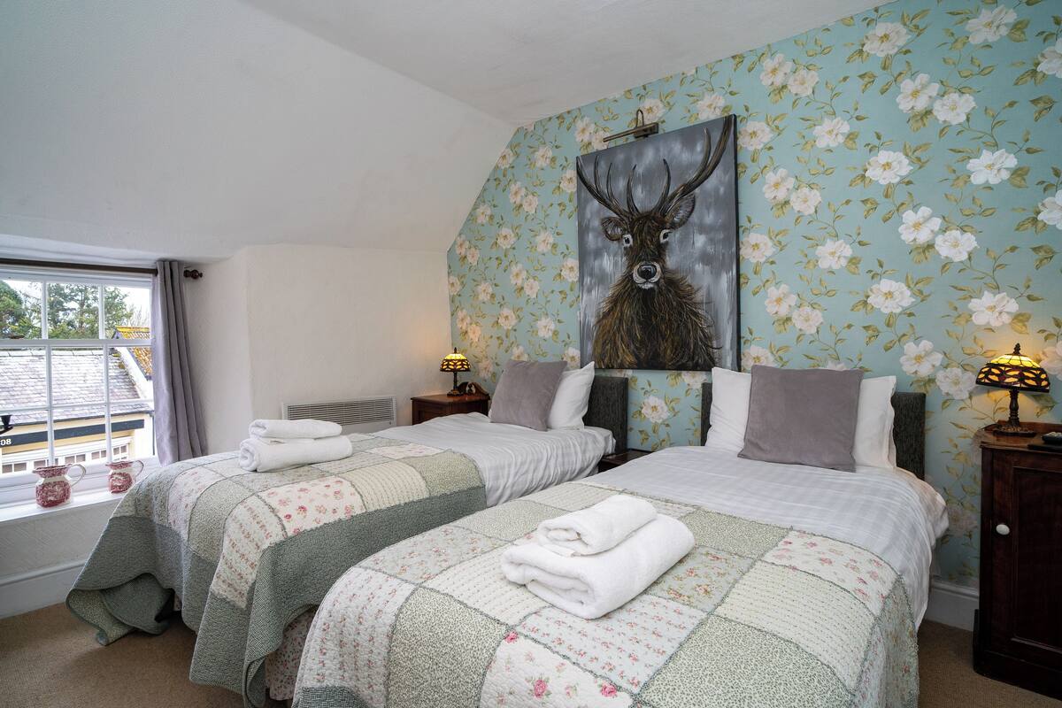 A bedroom with two beds, floral wallpaper, a deer painting, and a window with curtains.