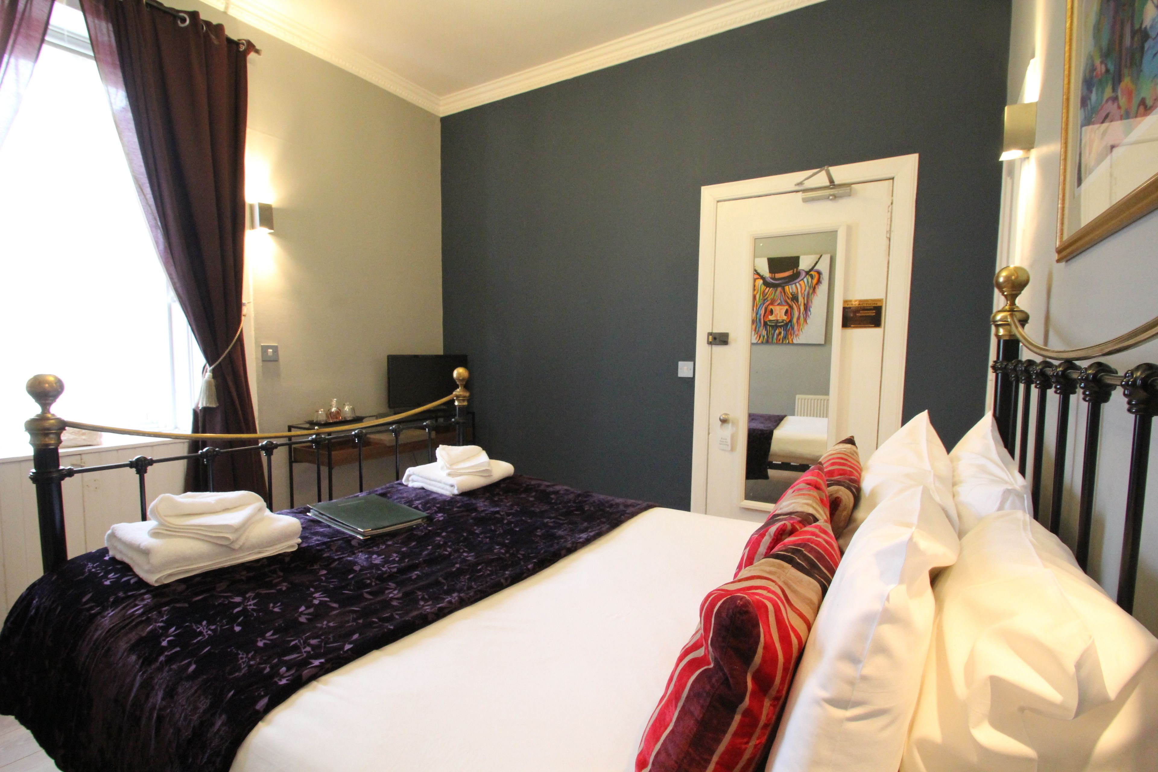 Ein Hotelzimmer mit einem Bett, einem Schreibtisch und einem Sessel. Es gibt ein Fenster mit Vorhängen, eine Tür und ein Bild an der Wand.