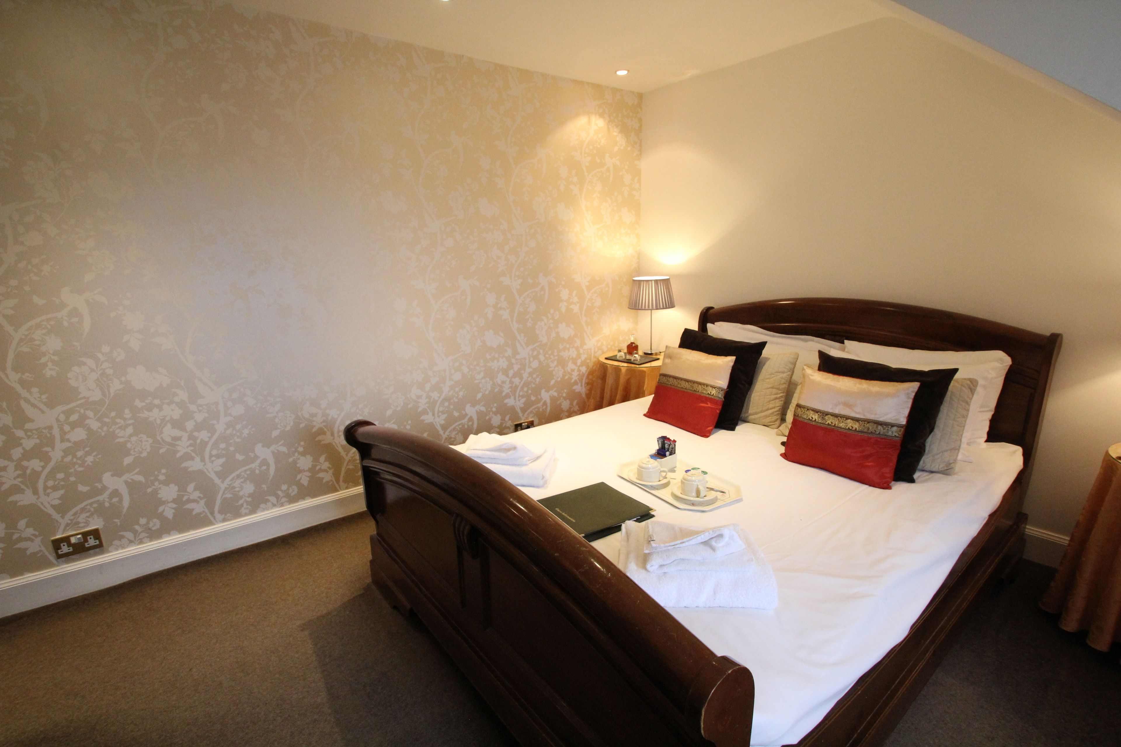 A neatly made bed with pillows, a bedside table with a lamp, and a floral wallpaper pattern.