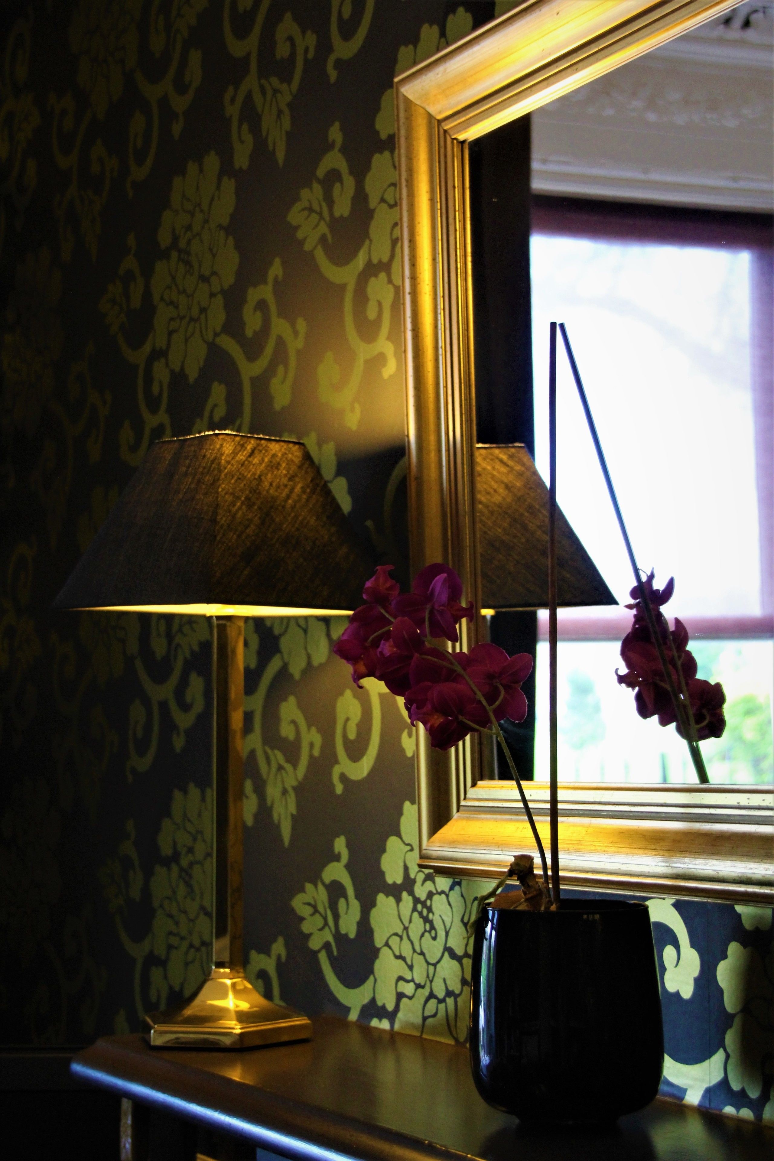 A lamp with a dark shade, a vase with purple flowers, and a mirror reflecting a window with daylight.