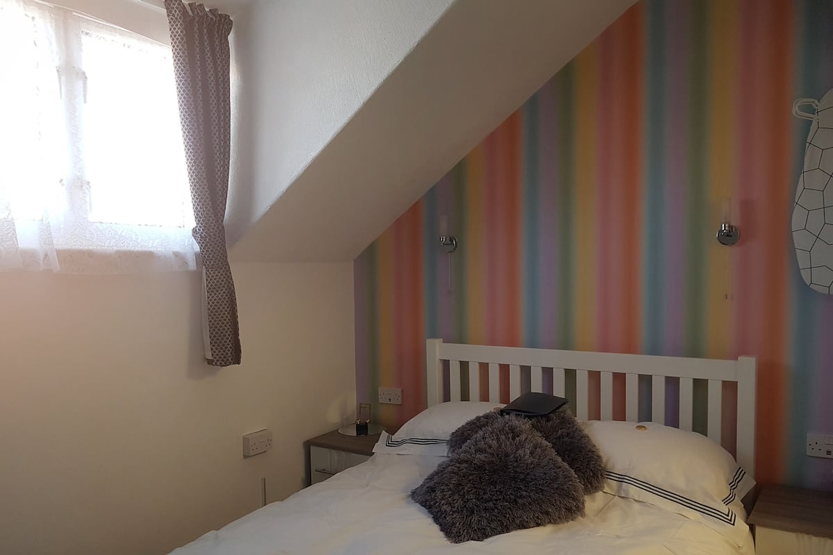 A bedroom with a white bed, a rainbow-striped wall, a white headboard, and a window with a curtain.