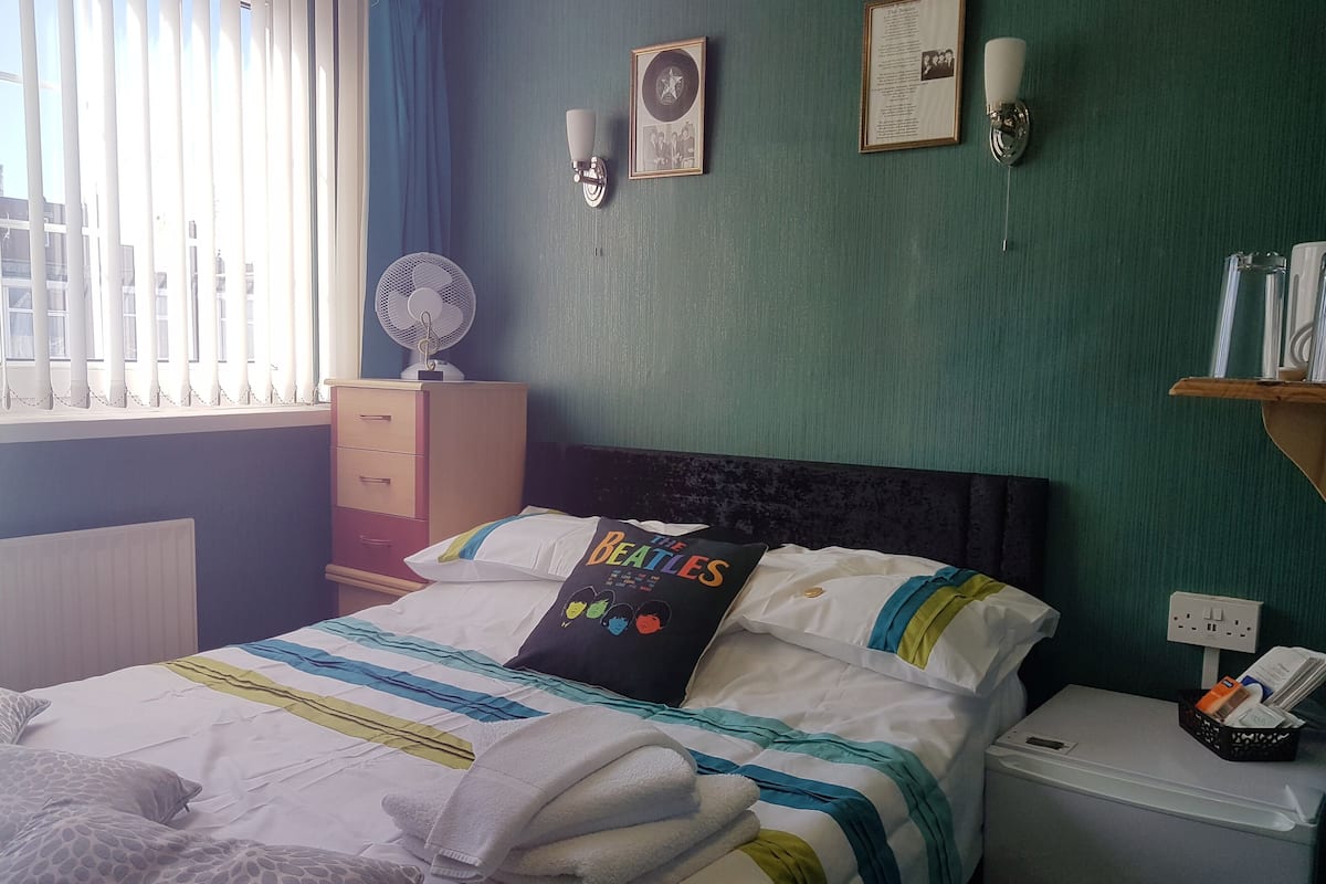 A neatly made bed with a 'The Beatles' pillow, a bedside fan, and a small bedside table with a lamp and a phone.