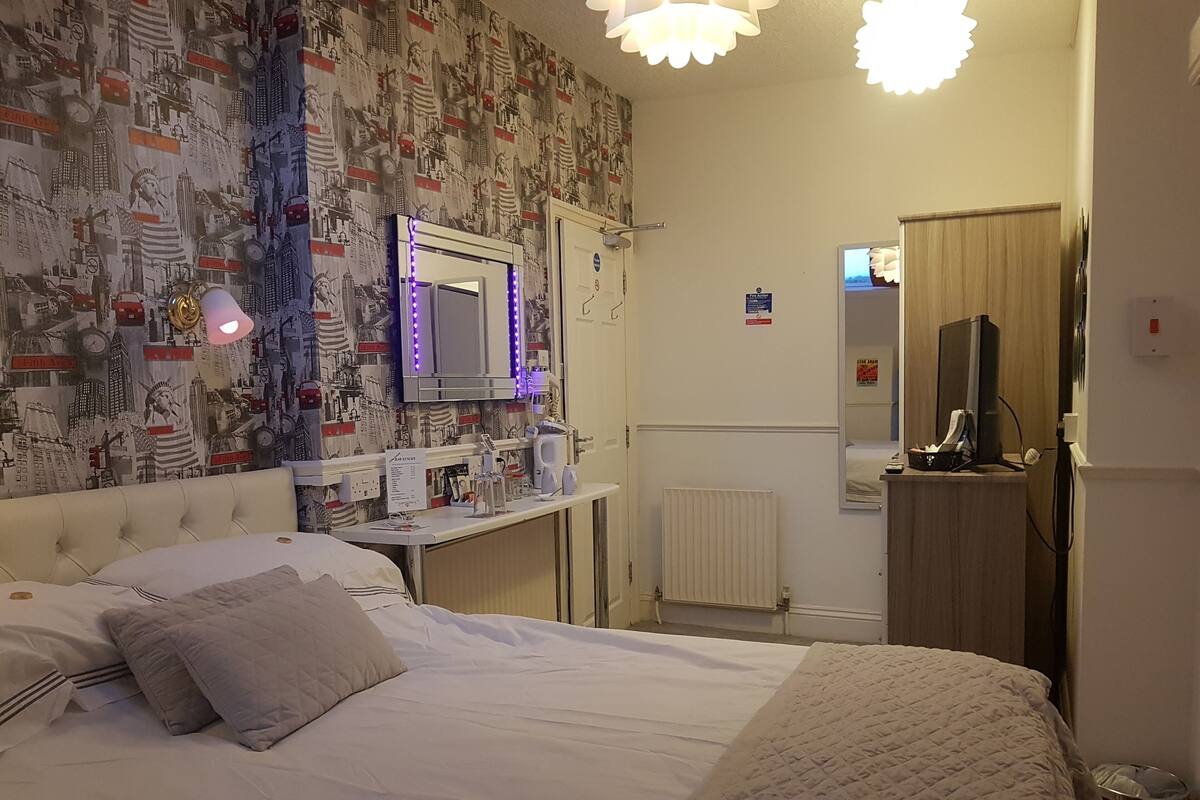 A hotel room with a bed, a vanity with a mirror, a desk with a computer, and a wall with a cityscape wallpaper.