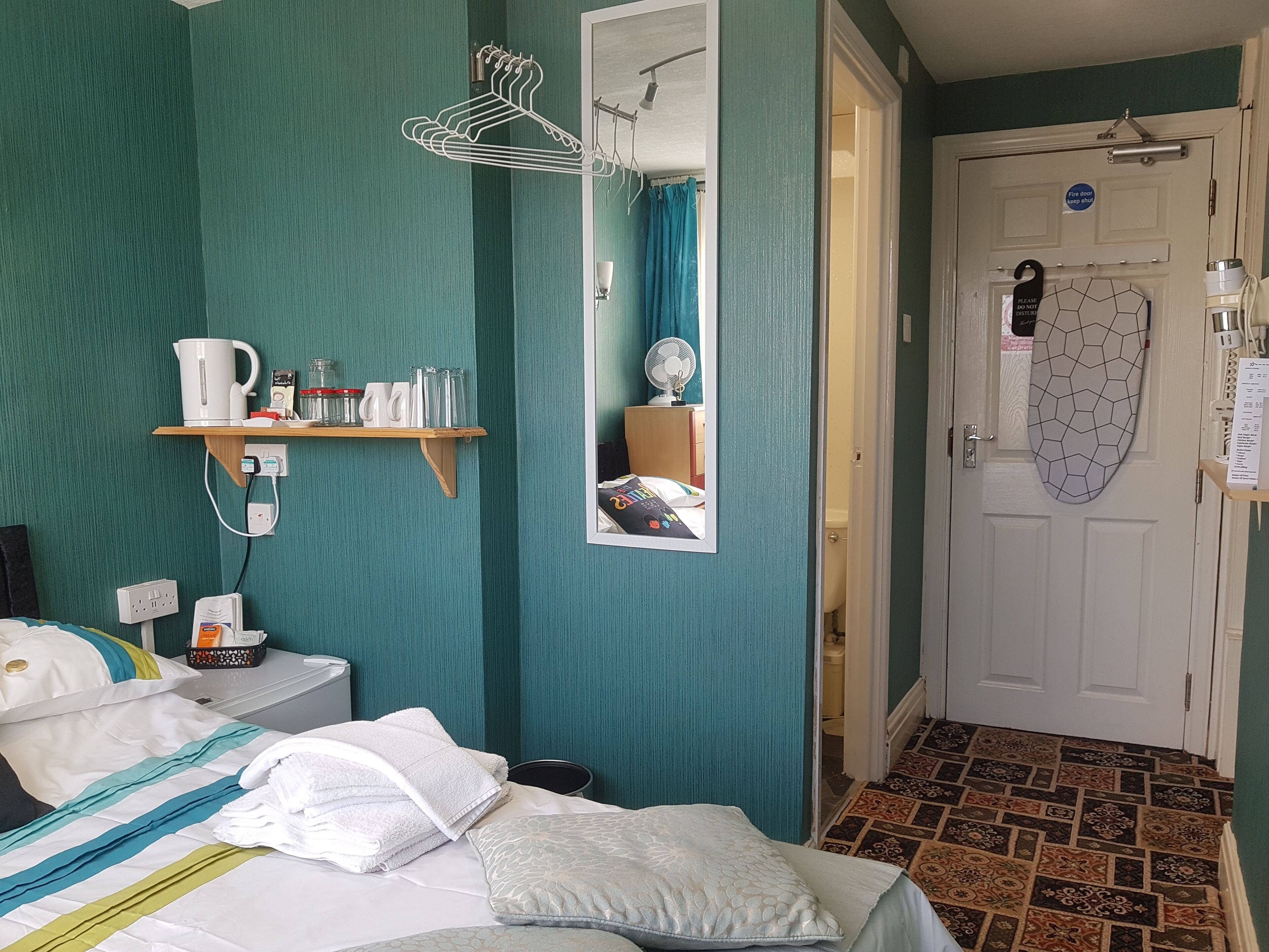A hotel room with teal walls, a bed with white and teal bedding, a bedside table with a kettle and mugs, a mirror, and a door with a fire safety sign.