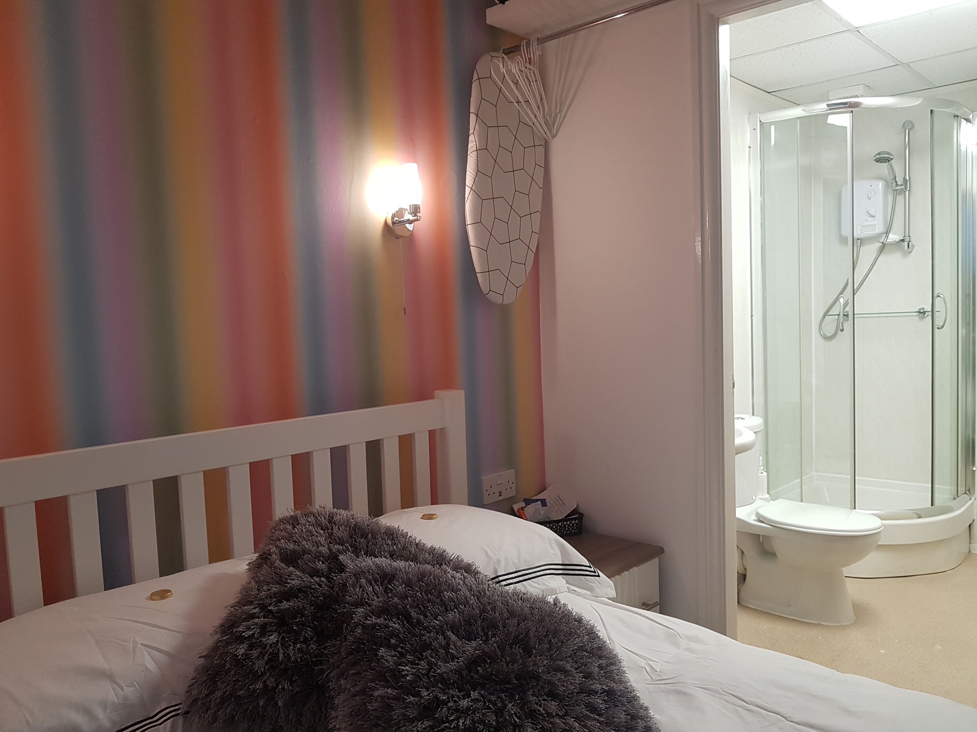 A bedroom with a bed, a bedside table, a shower area with a glass door, and a colorful striped wall.