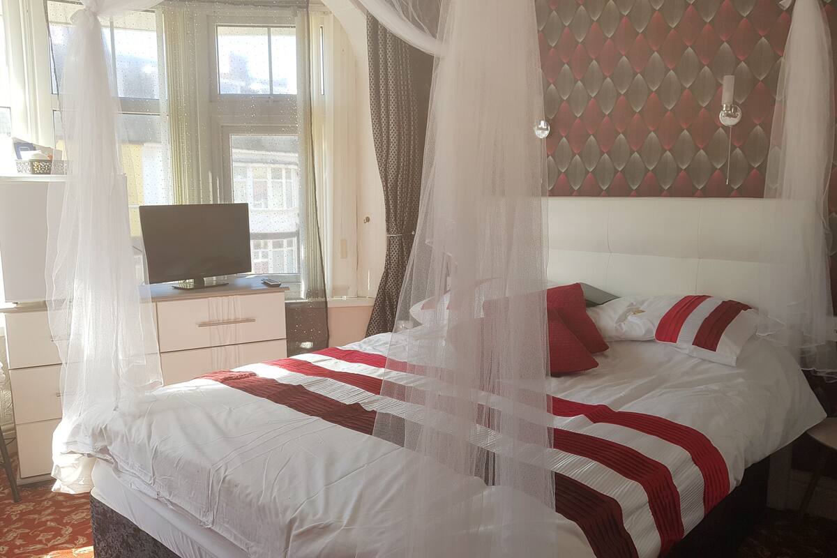 A bedroom with a four-poster bed, a television on a dresser, and a window with curtains.