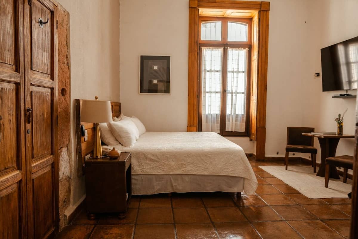 Une chambre à coucher comprenant un lit, une table de chevet, une lampe, une armoire en bois, une fenêtre avec des rideaux et un téléviseur fixé au mur.