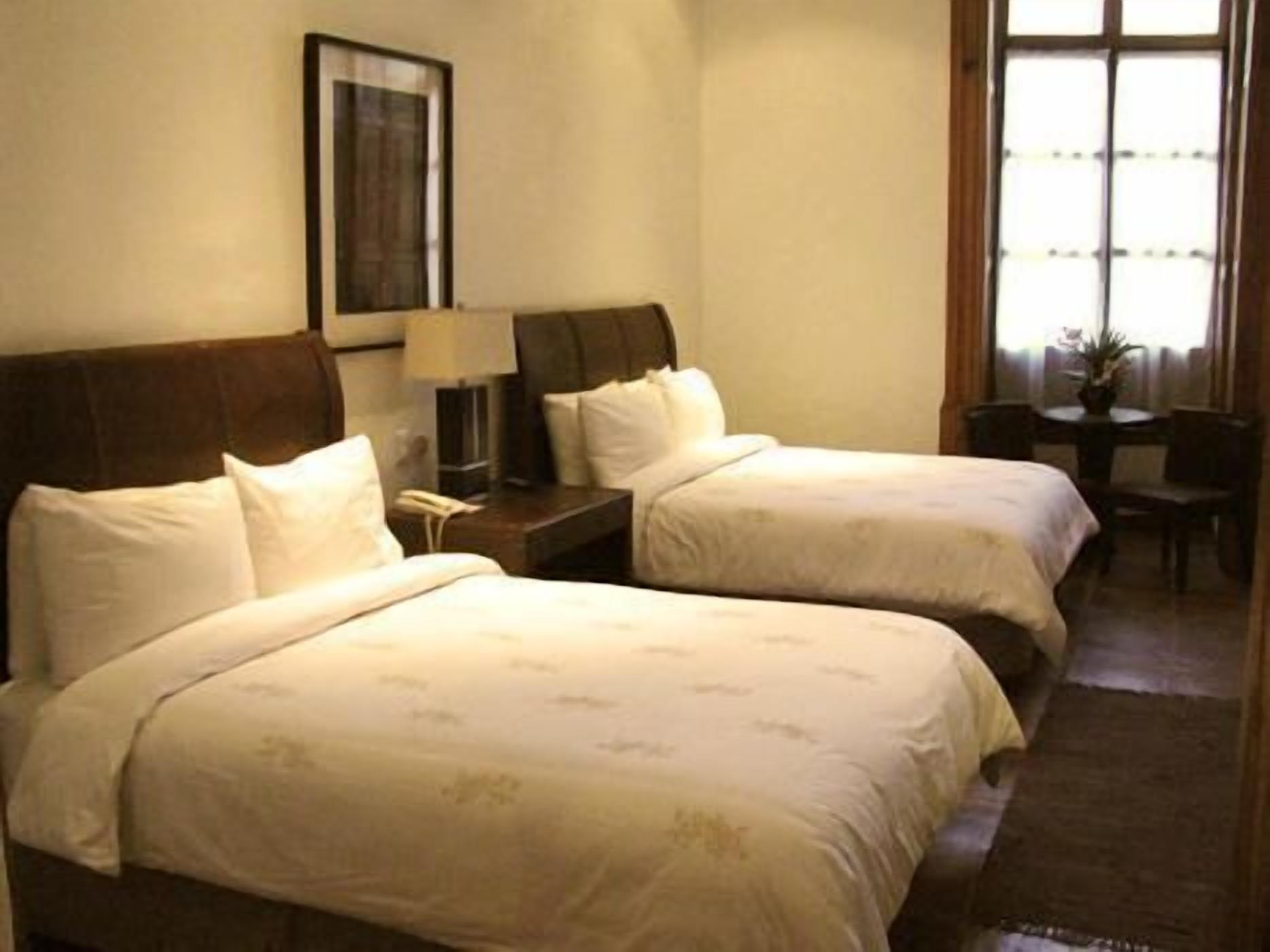 Habitación de hotel con dos camas, una mesita de noche con lámpara, una pequeña mesa con silla y una ventana con cortinas.