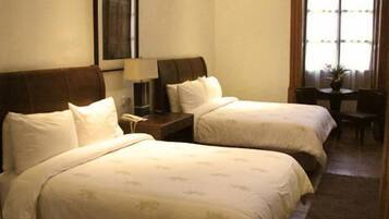 Habitación de hotel con dos camas, una mesita de noche con lámpara, una pequeña mesa con silla y una ventana con cortinas.