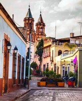 Una calle empedrada con edificios históricos, una iglesia con un campanario y un pequeño parque.