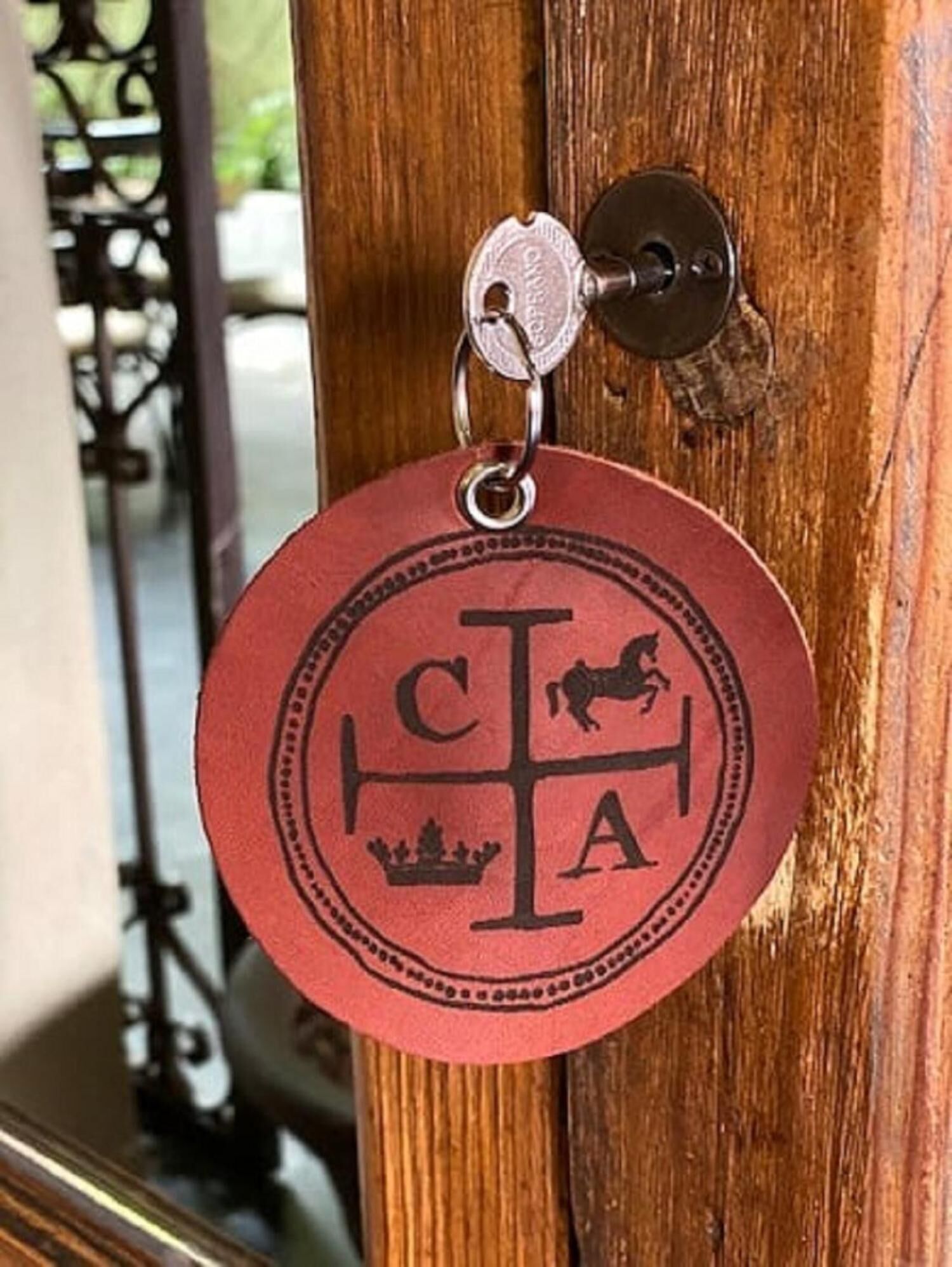 Um porta-chaves vermelho circular com um brasão, preso a um argol, pendurado numa porta de madeira.