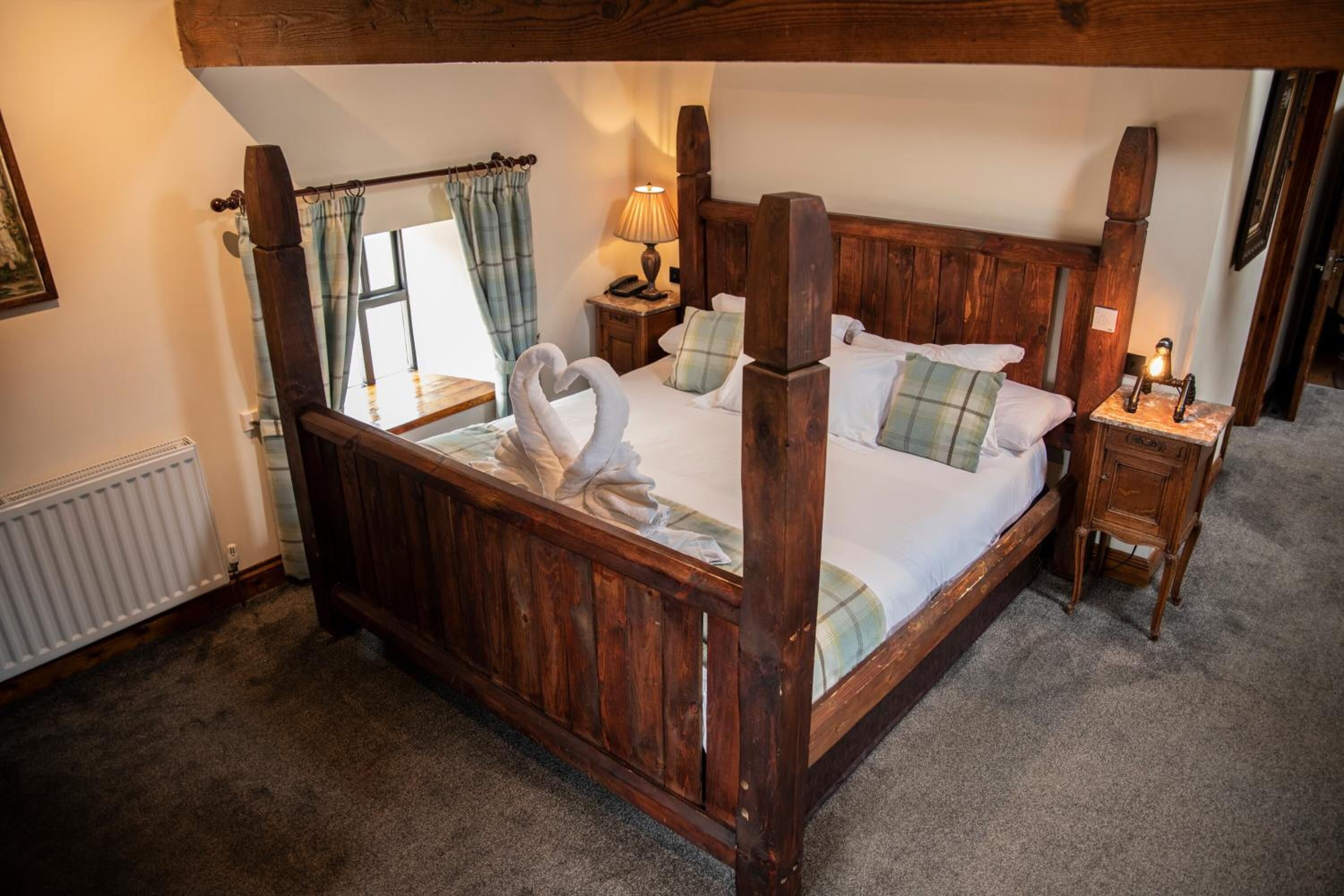 A four-poster bed with a wooden frame, a swan-shaped towel, and plaid bedding. There is a nightstand with a lamp and a telephone.