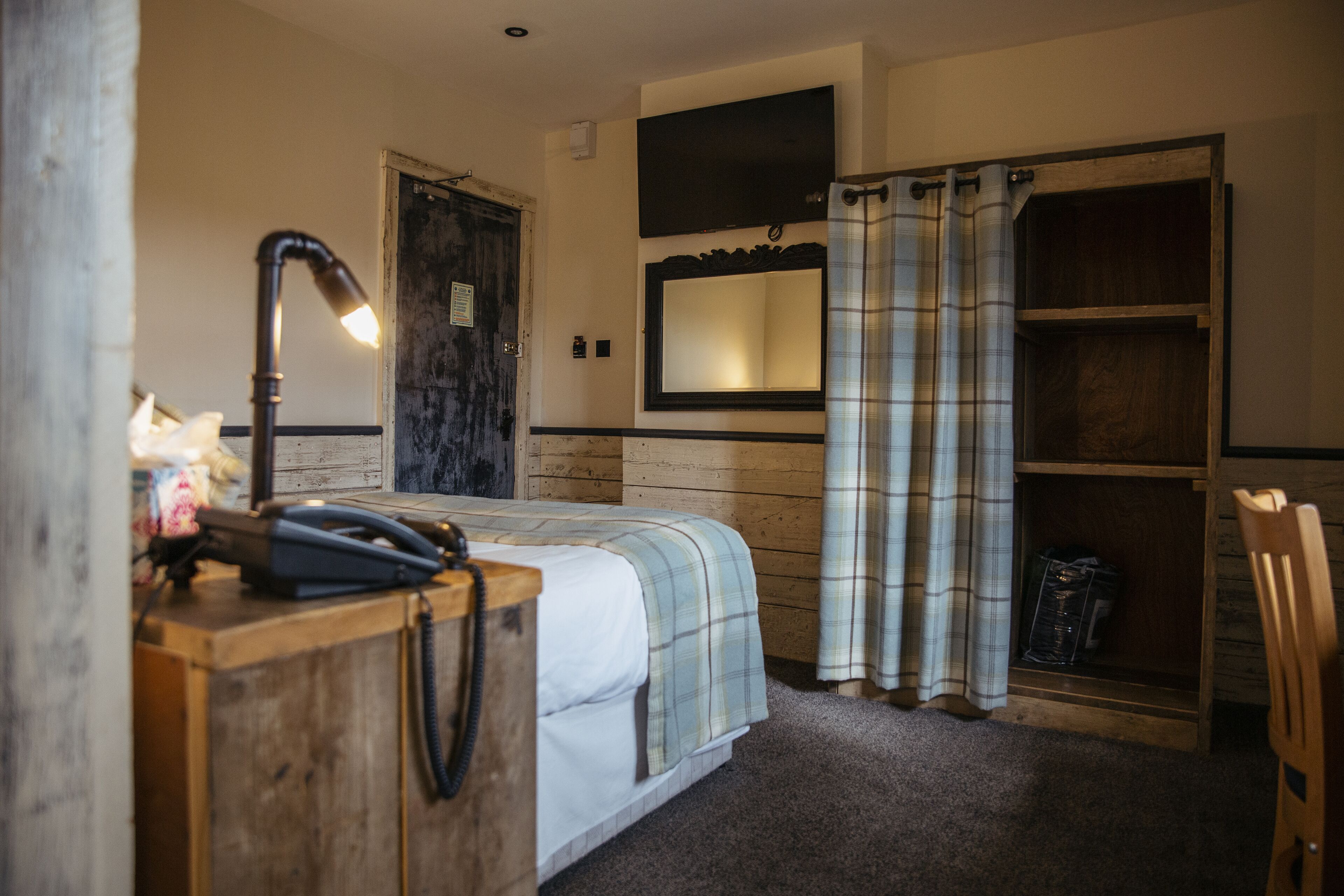 A bedroom with a bed, a desk with a lamp, a mirror, a wardrobe, and a shower area.