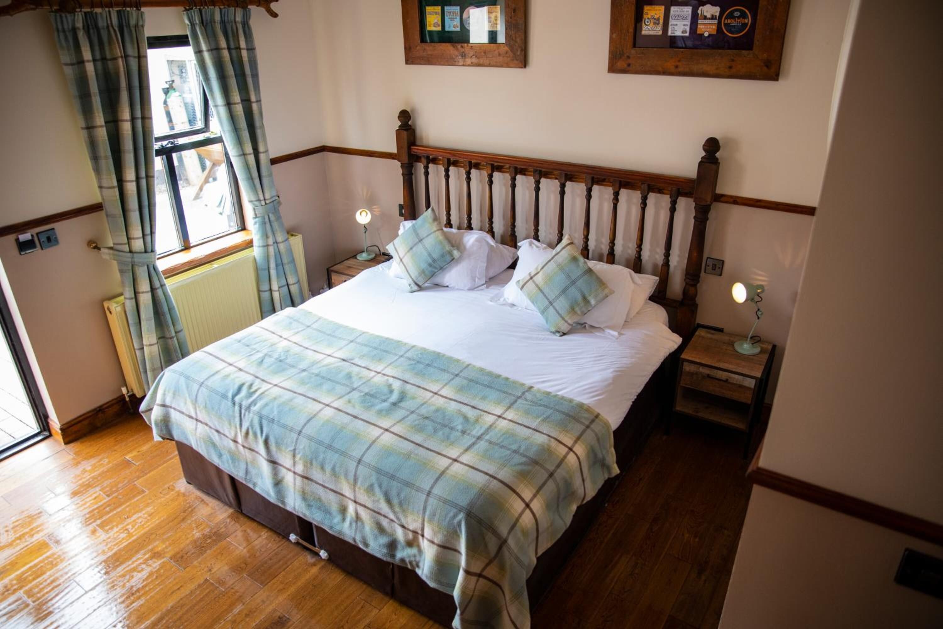 A bedroom with a wooden bed, plaid bedding, two bedside tables with lamps, a window with curtains, and a radiator.