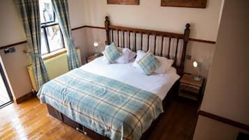 A bedroom with a wooden bed, plaid bedding, two bedside tables with lamps, a window with curtains, and a radiator.