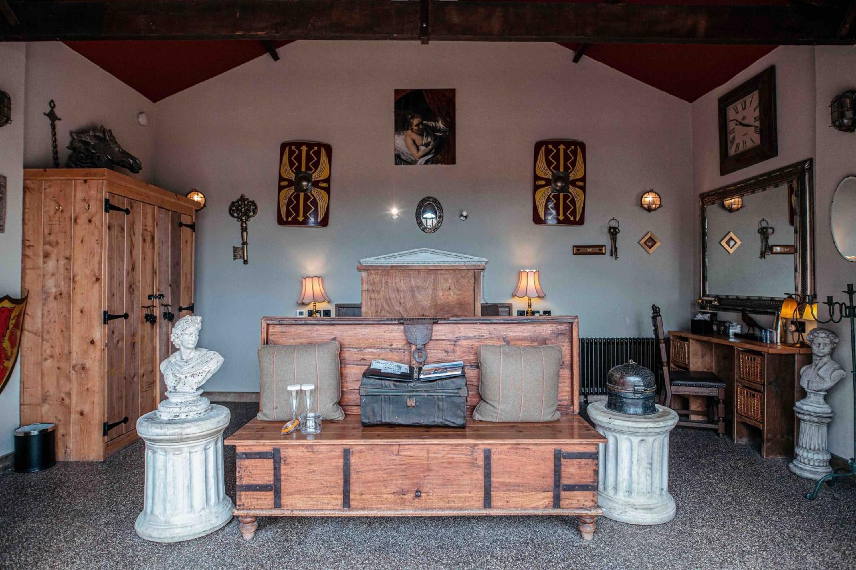 Een kamer met een houten kast, een borstbeeld op een voetstuk, een houten bank met kussens en een spiegel aan de muur.