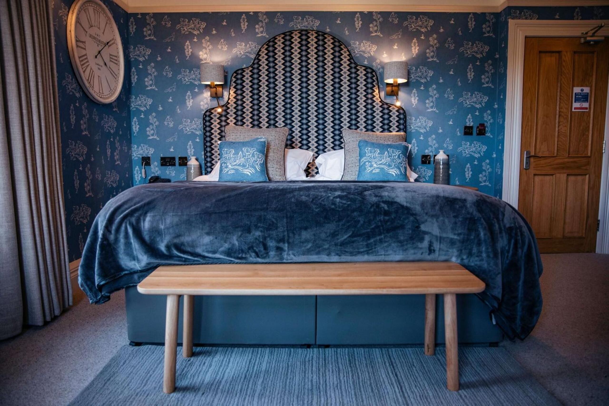 A bedroom with a patterned headboard, bedside lamps, a clock, and a bench.