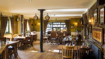 Un restaurante con mesas y sillas de madera, un reloj grande y una pared de piedra con cuadros enmarcados.
