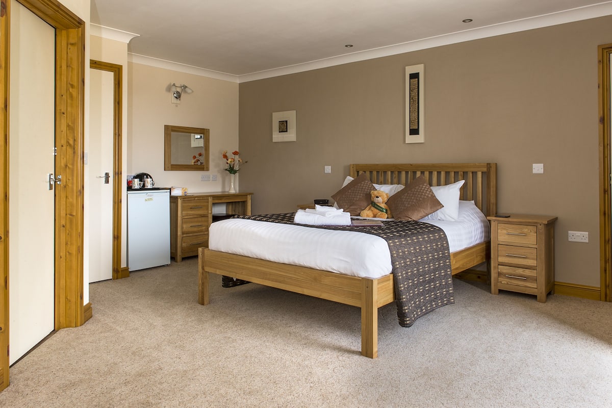A bedroom with a wooden bed, bedside tables, a dresser, a small desk, and a refrigerator.